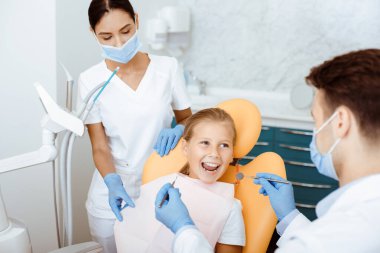 Hızlı, acısız ve modern muayene ve ağız boşluğu tedavisi. Gülümseyen küçük kız tıbbi sandalyede ağzını açar ve beyaz önlüklü, koruyucu maskeli ve eldivenli hemşireye bakar.