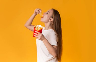 Hile Günü Konsepti. Aç Milenyum Kızı Fransız kızartması yiyor, Sarı Stüdyo 'nun arka planında duruyor, bedava manzarası var.