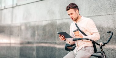Genç bir adam tesadüfen bisikletinde oturuyor, elinde tabletle kahve fincanı tutuyor. Gündüz vakti modern bir şehir ortamında odaklanmış ve rahat görünüyor.