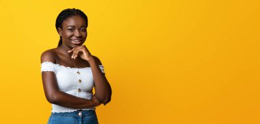 Sarı arka planda kendine güvenen siyah kadın, komik saçlı, şık bluzlu, kot pantolonlu, çenesine dokunan ve kameraya bakan Afrikalı Afrikalı Amerikalı kadın.