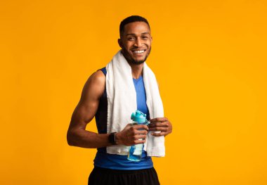 Susuz kalma. Boynunda havluyla sarı stüdyo arka planında su şişesi tutan kaslı zenci adam portresi.