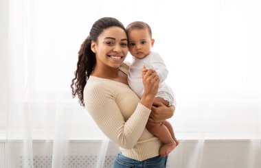 Afrika anne ve bebek portresi. Mutlu Siyah Anne, sevimli bebeğini kucağında tutarak içeride pencerenin yanında kameraya gülümsüyor. Neşeli Annelik Anları Kavramı
