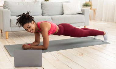 Siyah Sporcu Kadın kapalı alanda çalışırken dizüstü bilgisayarda Plank Egzersizi Yapıyor. Evde Eğitim Konsepti