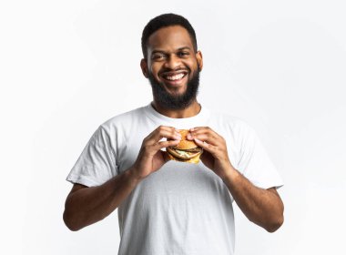 Abur cubur. Stüdyoda beyaz arka planda durup kameraya gülümseyen mutlu siyah adam hamburgeri tutuyor. Beslenme, aşırı yeme alışkanlığı ve sağlıksız yeme anlayışı