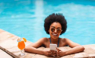 Neşeli siyah kız ferahlatıcı yaz kokteyli ve akıllı telefon ile güneşli bir günde havuzda dinleniyor, boşluğu kopyalıyor.