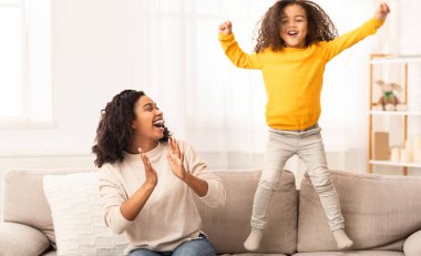 Aile eğlencesi. Mutlu siyah anne ve kızı eğleniyor, çocuk evde kanepeye zıplıyor.