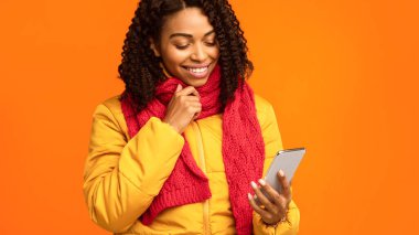 Cep telefonlu Siyah Kadın Çevrimiçi Mesajları Okuyor, Sarı Stüdyo Arkaplanı Üzerinde Cep Telefonu Bekliyor, Kış Ceketi Giyiyor. Akıllı Telefon Uygulama Konsepti. Kırpılmış, Boşluğu Kopyala