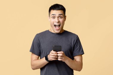 İnternette ya da büyük miktarda para kazanmanın harika haberleri var. Mutlu, heyecanlı, şoke edici, akıllı saati ve ağzı açık genç bir adam. Akıllı telefon, açık kumlu arka planda izole edilmiş, boş alan.
