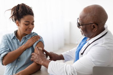 Antiviral aşı, bağışıklık sistemi. Sağlık merkezinde enjeksiyondan sonra hastaların omzuna alçı bandaj yapıştıran olgun bir doktor. Hamile siyahi kadın Covid önleme aşısı oluyor.