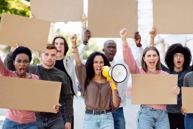 Megafonlu melez bir kadın sokakta boş pankartlarla bir grup göstericiyi yönetiyor. İnsan haklarını protesto eden uluslararası bir grup insan, boş tahtaları gösteriyor