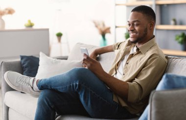 Harika Mobil Uygulama. Mutlu Afro-Amerikalı Adam telefon mesajlaşması yapıyor ve internette geziniyor. Evdeki koltukta oturuyor, yandan bakıyor. Smartphone 'da Siyah Erkek Ağı Sosyal Medya Uygulaması Kullanıyor