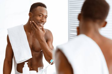 Erkek Cilt Bakımı. İçerideki banyoda aynanın yanında duran cilt bakımı yapan neşeli Afrikalı adam pürüzsüz yüze dokunuyor. Erkek Güzelliği ve Kişisel Bakım Rutini. Seçici Odaklanma