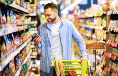 Tüketim Konsepti. Gülümseyen Sakallı Adam 'ın portresi markette alışveriş arabasıyla marketten yiyecek alıyor koridorda yürüyor ve raflarda duruyor, elinde cam kavanoz sos tutuyor, etiketi kontrol ediyor.