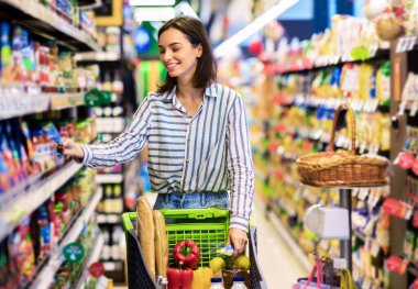 Her gün alışveriş konsepti. Gülümseyen Genç Ev Kadını Markette Koridorlar Arasında Trolley Arabası İtiyor. Pozitif kadın yemek alıyor, raftan ürün alıyor. Tüketim ve Yaşam Tarzı