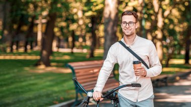 Kahve alıp işe gitmek için. Elinde bardak ve gözlüklerle çekici, mutlu bir adam parka bisikletle gider.