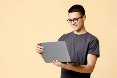 Modern uzaktan haberleşme devrede, evde kal. Gözlüklü gülümseyen genç öğrenci dizüstü bilgisayara bakar ve proje üzerinde çalışır, bej arka planda izole edilmiş, stüdyo çekimi