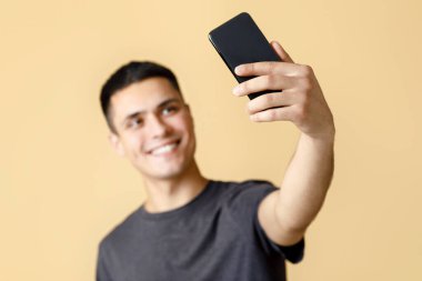 Happy blogger sosyal medya için video veya fotoğraf yayınlıyor. Kahverengi tişörtlü gülümseyen Z nesli, selfie, bulanık ve akıllı telefona odaklı, bej arka planda izole edilmiş, serbest alan yaratıyor.