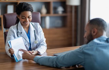 Smiling young black woman doctor showing middle eastern man patient treatment plan, arab guy attending therapist at newest private clinic, copy space, modern healthcare concept