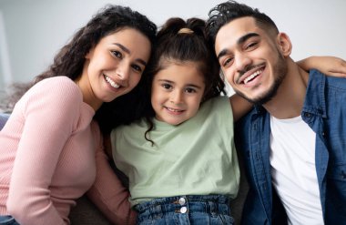 Üç çocuklu mutlu Arapça aile portresi, neşeli Orta Doğulu ebeveynler, küçük çocuklarıyla evde eğlenirken kamerada poz veriyorlar, birlikte koltukta dinleniyorlar.