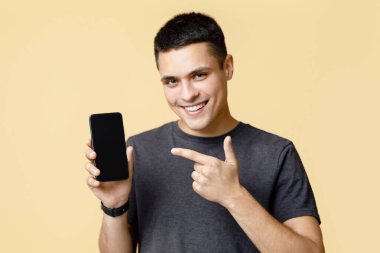 Heyecanlı kazanan adam iyi haberler aldıktan sonra cep telefonu gösterisi yapıyor. Gülümseyen genç adam, boş ekranlı akıllı telefonu işaret ediyor, iyi bir cihaz tavsiye ediyor, bej arka planda izole edilmiş.