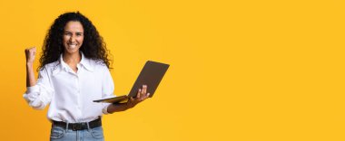 Öforik Esmer Kadın Dizüstü bilgisayarla başarıyı kutluyor, yumruğunu kaldırıyor, Mutlu Milenyum Kadın Tutan Bilgisayar, Zafer ve Başarı, Sarı Stüdyo Arkaplanında Durma, Kopya Boşluğu