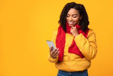 Akıllı telefonlu, cep telefonu haberleri ve mesajları okuyan, elinde telefon, sarı stüdyo geçmişinde mesajlaşan, kışlık giysiler giyen Afro-Amerikan bayan. Cep telefonu kullanıcı uygulaması kullanıyor. Boş Alan
