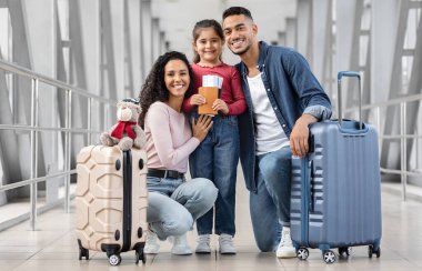 Havalimanı terminalinde üç poz veren mutlu orta doğulu aile, neşeli Arap ebeveynler ve küçük şirin kızlarının bavulları, pasaportları, biletleri ve kameralara gülümseyen, seyahat etmeye hazır.