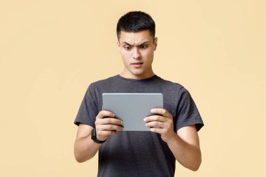 Harika haberler, şok edici mesajlar, problemler ve oyunda kaybetmek. Şaşırmış genç erkek öğrenci ya da blog yazarı şaşkınlıkla tablete bakıyor, bej arka planda izole edilmiş, stüdyo çekimi, boş alan