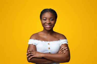 Portrait of confident young african american lady standing with folded arms over yellow background, succesful millennial black woman in stylish clothes looking and smiling at camera, free space