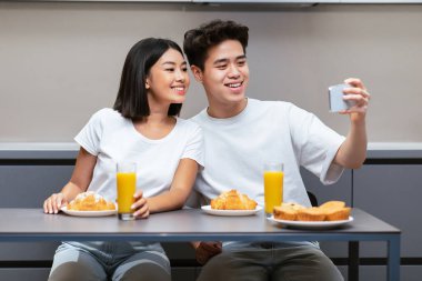 Mutlu Koreli koca, evde mutfakta otururken karısıyla selfie çekiyor. Asyalı aile, hafta sonu sabah kahvaltısında portakal suyu içip hamur işi yiyor.