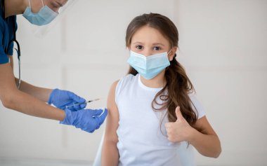 Sağlık merkezinden kovid 19 aşısı alırken yüz maskeli küçük kız başparmağını kaldırıyor. Koronavirüs aşısını onaylayan sevimli çocuk küresel virüse karşı korunuyor.