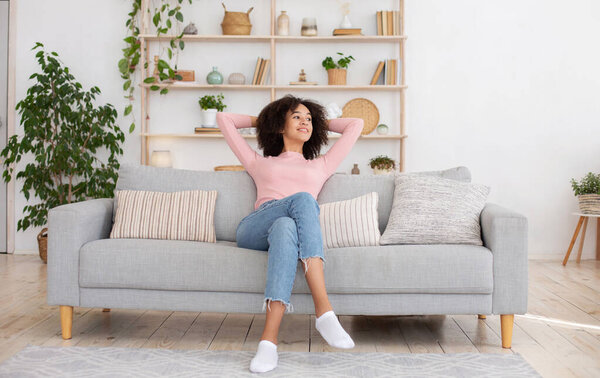 Чистота в квартире, новый дом и положительные эмоции. Улыбающаяся молодая африканская американка наслаждается тишиной, бросает руки за голову и смотрит в сторону, сидит на диване в интерьере гостиной