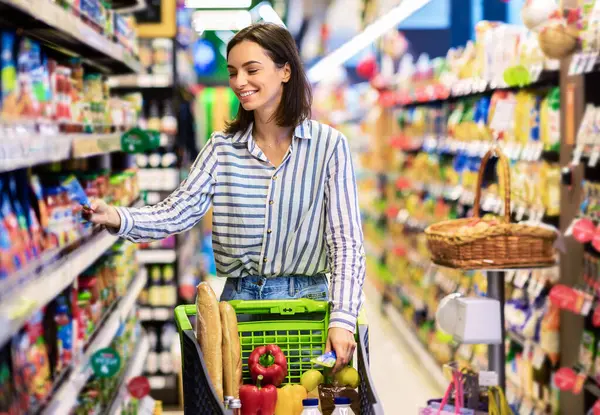 Her gün alışveriş konsepti. Gülümseyen Genç Ev Kadını Markette Koridorlar Arasında Trolley Arabası İtiyor. Pozitif kadın yemek alıyor, raftan ürün alıyor. Tüketim ve Yaşam Tarzı