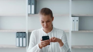 Genç bir iş kadını, çağdaş bir ofis ortamında akıllı telefonuna odaklanır. Beyaz bir bluz giyiyor ve işine dalmış, kurumsal bir hayat tarzı sergiliyor..