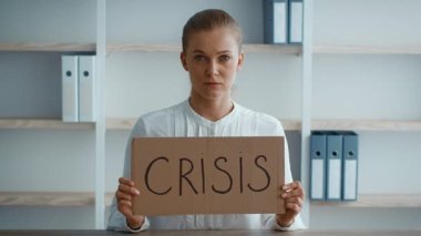 Modern bir ofiste masa başında oturan genç profesyonel bir kadın elinde CRISIS yazan bir karton tabela tutuyor. Odaklanmış ve ciddi görünüyor, kurumsal bir atmosferi somutlaştırıyor..