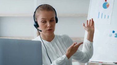Beyaz bluzlu genç bir kadın modern bir ofisteki kulaklıkla aktif olarak iş arkadaşlarıyla takılıyor. Bir sunuma doğru el kol hareketi yapıyor, şirket verilerini ve grafiklerini gösteriyor..