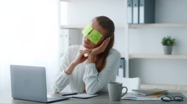 Kurumsal ofis ortamında oturan genç bir kadın, bir an rahatlarken gözlerini kapatmak için yapışkan notlar kullanıyor. Düşünceli görünüyor ve sakin atmosferden hoşlanıyor..