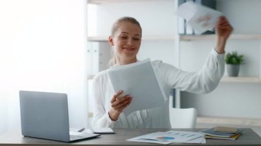 Genç bir kadın ofisinde belgeleri incelerken sevinçten havalara uçuyor. Profesyonel giyinmiş ve modern ofis malzemeleriyle çevrili. Canlı bir şirket hayatı sergiliyor..