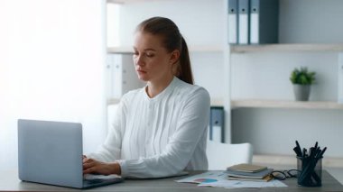 Modern ofis ortamında dizüstü bilgisayarına odaklanmış genç bir kadın. Etrafı defterler ve kağıtlarla çevriliyken işiyle meşgul görünüyor..