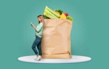 Müşterinin biri telefonunu, içi taze ürünlerle dolu büyük bir kesekağıdına yaslanırken kontrol ediyor. Görüntü, market alışverişinin ve teslimatın çağdaş bir ortamda yeni normalini vurguluyor.