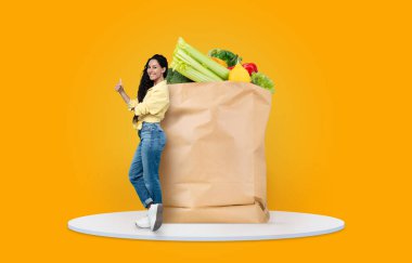 Neşeli bir müşteri taze sebze ve meyvelerle dolu büyük bir poşetin yanında dururken başparmağını kaldırır. Geçerli arka plan alışveriş deneyimini güçlendirir.