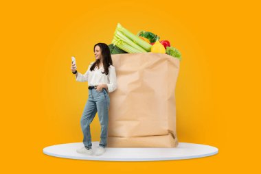 Bir müşteri, içi taze yiyecek dolu büyük kahverengi bir kese kağıdının yanında mutlu bir şekilde durur. Akıllı telefonlarıyla meşgul oluyorlar, muhtemelen teslimat için erzak sipariş ediyorlar..