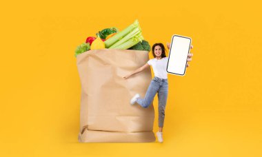 Bir müşteri, içi taze yiyeceklerle dolu büyük bir kese kağıdının yanında dururken gülümser. Elinde bir akıllı telefon var, online alışverişin kolaylığını gösteriyor. Bu, modern market alışverişini temsil ediyor..