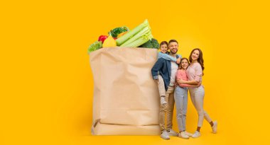 Bir aile, içi taze yiyeceklerle dolu büyük bir kesekağıdıyla mutlu mesut ayakta duruyor. Taze yemeğin tadını çıkarırken evde alışveriş yapmanın yeni normalliğini sembolize ediyorlar..