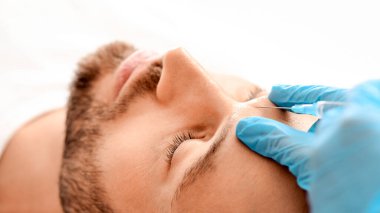 Klinikte güzellik iğnesi yaptıran sakallı adama yakın çekim. Kozmetik uzmanı, yakışıklı bir adam için yüz doldurma işlemi için mavi lastik eldivenleri teslim etti. Yüz bakımı, erkek kozmetoloji konsepti.