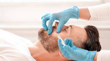 Estetik kliniğinde güzellik iğnesi yaptıran sakallı adamın yan görüntüsü. Estetik cerrah yakışıklı erkek yanaklarına yaşlanma önleyici enjeksiyon yapıyor. Erkek kozmetolojisi, estetik tıp konsepti