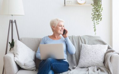 Evdeki koltukta oturan, dizüstü bilgisayarı olan ve telefonla konuşan modern mutlu yaşlı kadın.