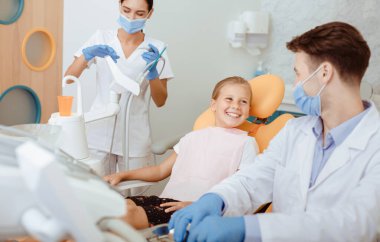 Takımla hızlı ve kolay diş tedavisi. Gülümseyen kız beyaz önlüklü, koruyucu maskeli ve eldivenli doktora bakıyor. Modern kliniğin içindeki dişçi ve hemşireye teşekkürler.