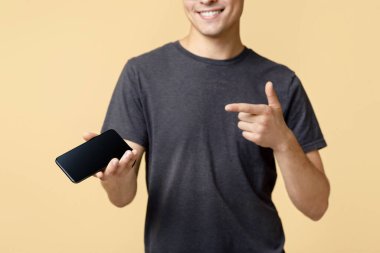 Cep telefonundan iyi bir teklif geldi. Kahverengi tişörtlü ve gözlüklü arkadaş canlısı genç bir adam, boş ekranlı akıllı telefonu işaret ediyor, bej arka planda izole edilmiş, stüdyo çekimi, boş alan.