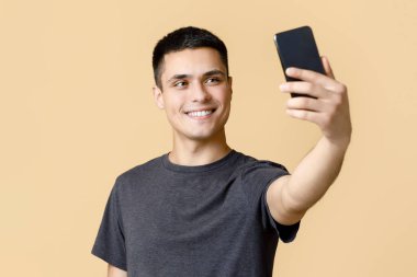 Blog için modern fotoğraf. Neşeli genç erkek öğrenci, sosyal ağlar için telefonda selfie çekiyor ve tavsiyelerle dolu bir video çekiyor, bej arka planda izole edilmiş, stüdyo çekimi, boş alan.